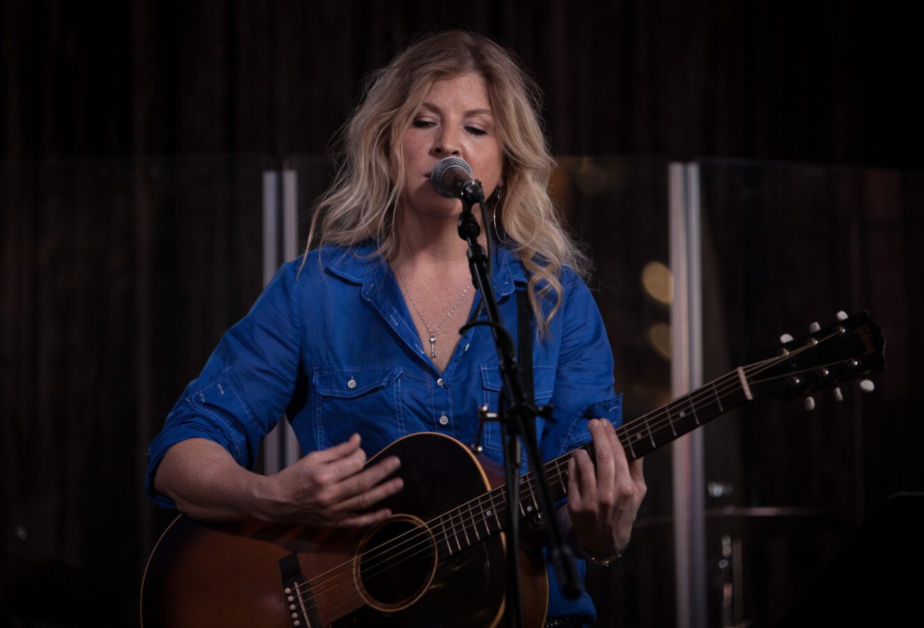 Singer-songwriter Amy Hailsatone strums an acoustic guitar while singing into a microphone onstage.