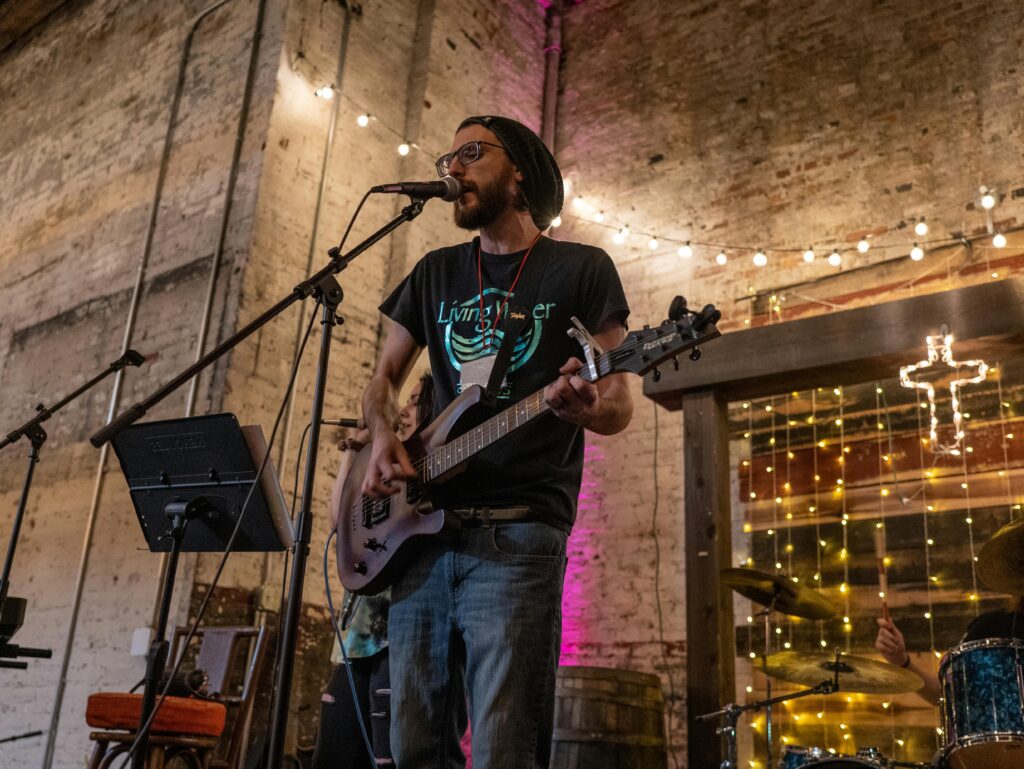 A thin man in a stocking hat plays guitar and sings into a mic on a stage with a brick wall and string lights in the background. 