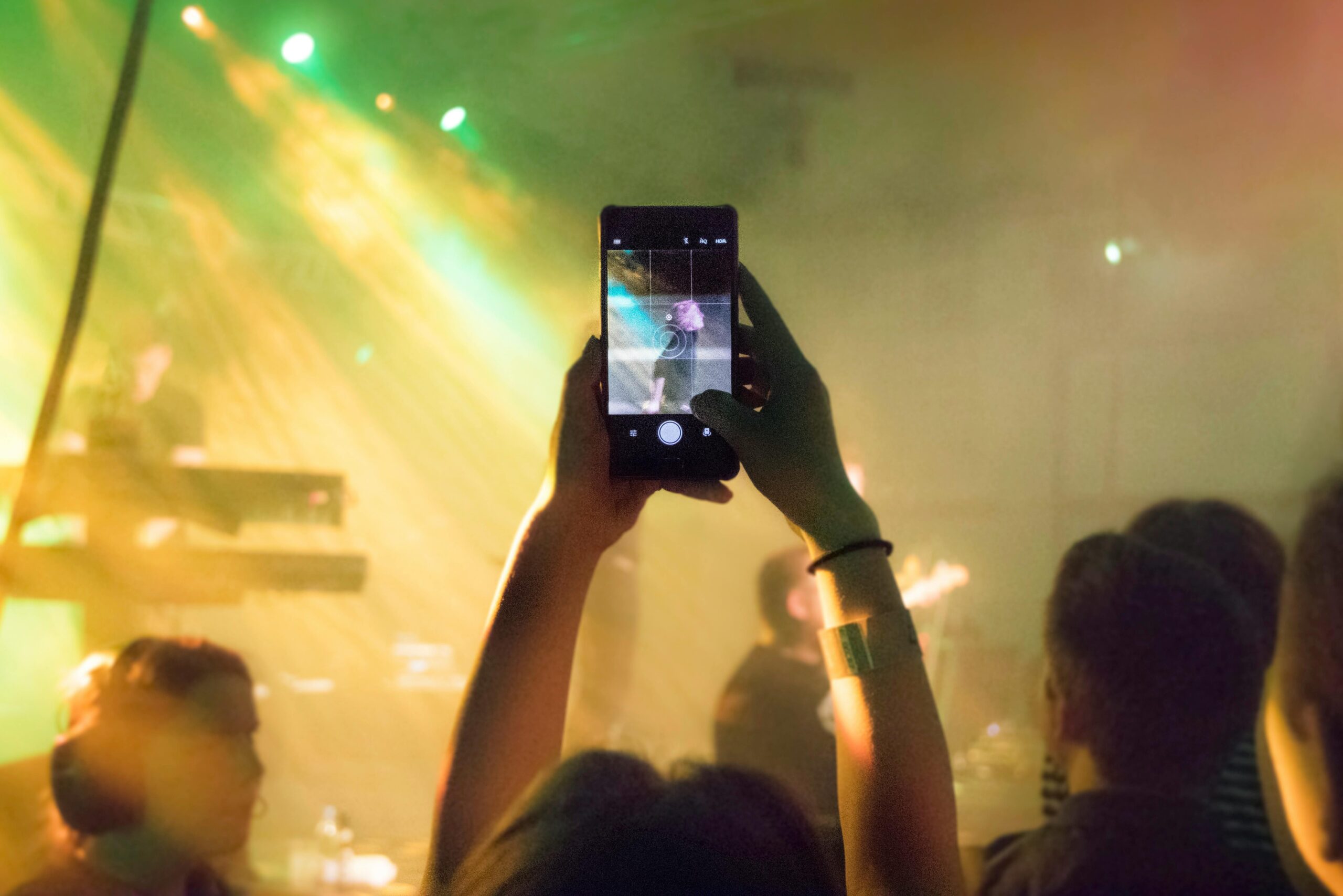 A concert goer holds their phone up to capture the musicians performing onstage awash in soft yellow beams of light.